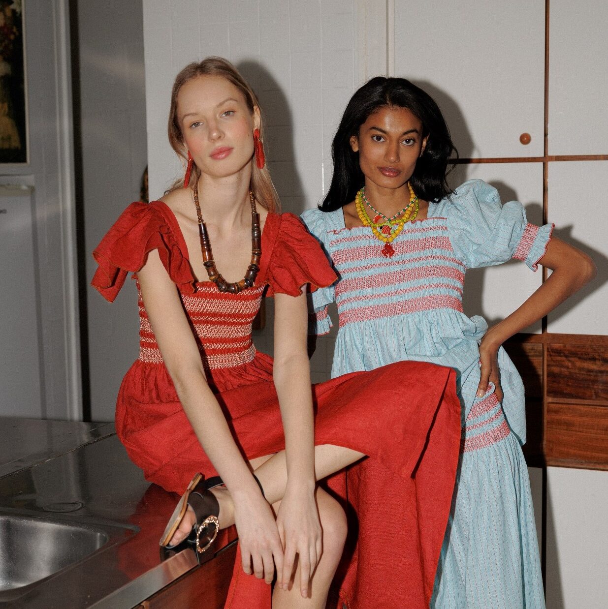 Dos mujeres posando con elegancia en la cocina, una con un vestido rojo con detalles de volantes y la otra con un vestido azul plisado y accesorios coloridos.