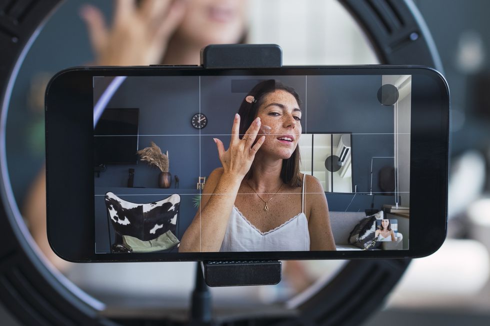 Mujer aplicando crema en su rostro mientras graba un video con su teléfono, capturada a través de un aro de luz.