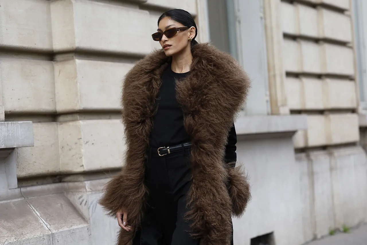 Mujer con abrigo de piel sintética marrón y gafas oscuras, posando con actitud en una calle urbana.