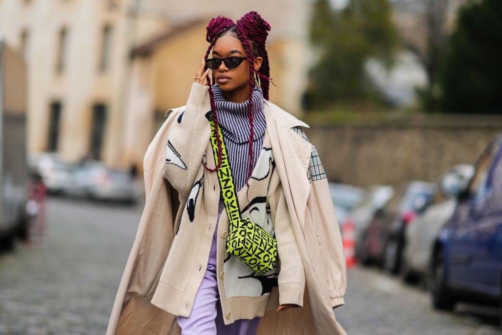 Mujer con trenzas rojas y gafas de sol, vistiendo un abrigo beige y un bolso con estampado neón mientras camina por la ciudad.