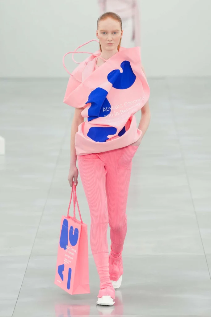 Propuesta de Issey Miyake en la Semana de la Moda de París 2025. Un conjunto en rosa vibrante con texturas plisadas y un top escultural con estampados gráficos en azul. El diseño, acompañado de una bolsa a juego, fusiona el arte conceptual con la moda contemporánea.