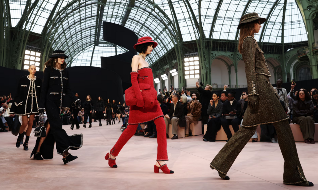 esfile de Chanel en París Fashion Week con modelos presentando la nueva colección en un escenario majestuoso. Destaca un conjunto de tweed rojo con guantes y bolso a juego, mientras otras modelos lucen icónicos trajes de la maison en negro y dorado.