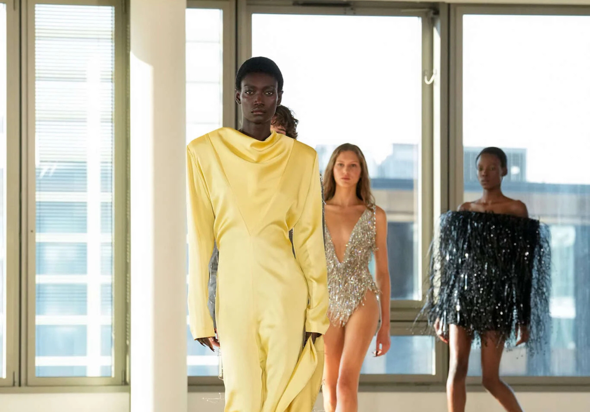 Modelos desfilando en la pasarela de Stella McCartney Otoño-Invierno en la semana de la moda de Paris 2025. En primer plano, un vestido estructurado en amarillo pastel con cuello drapeado, seguido de un body de cristales plateados y un mini vestido negro con texturas de flecos brillantes.