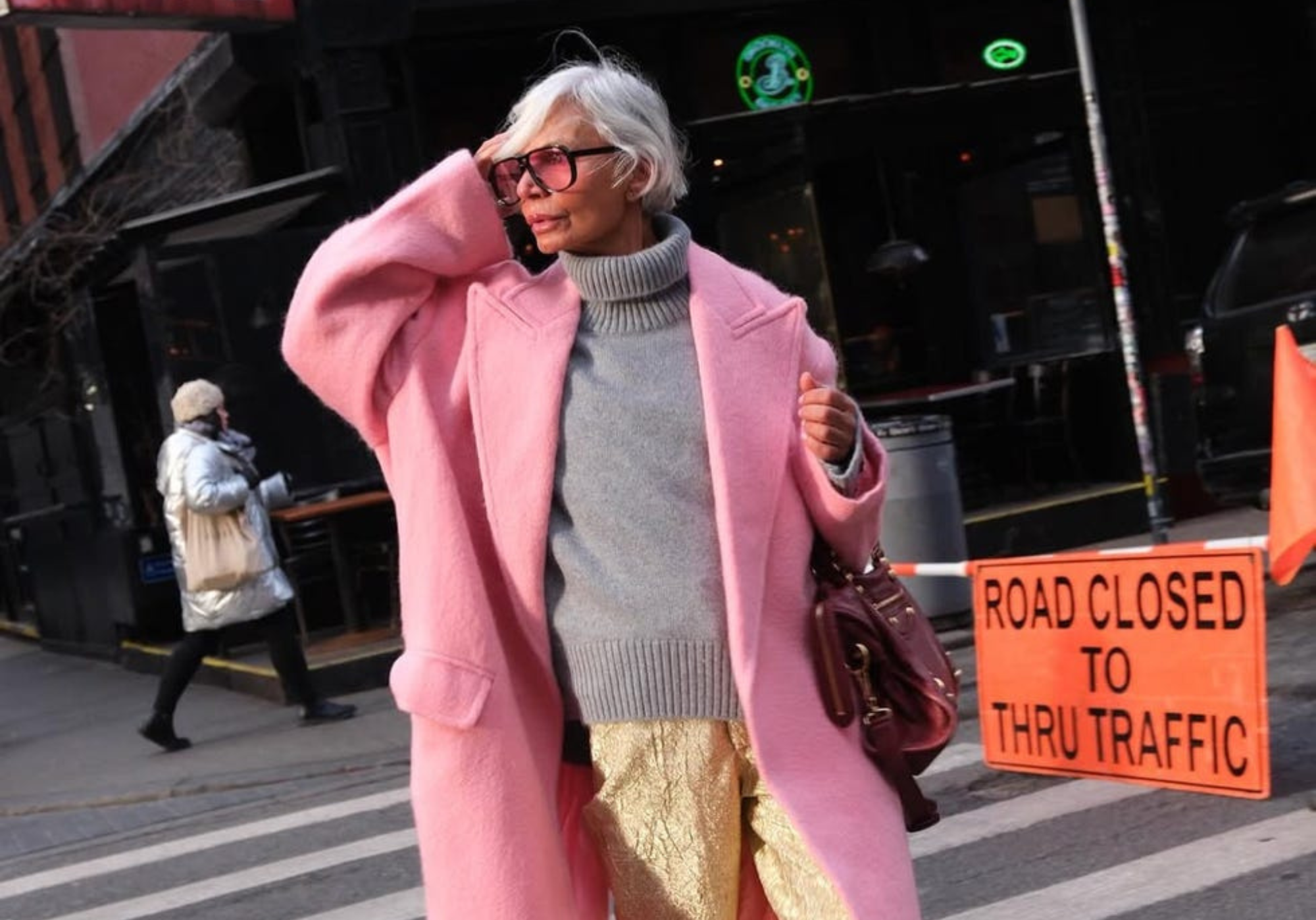 Mujer mayor con cabello blanco y gafas de sol, usando un abrigo rosa, suéter gris y pantalones dorados en la calle.