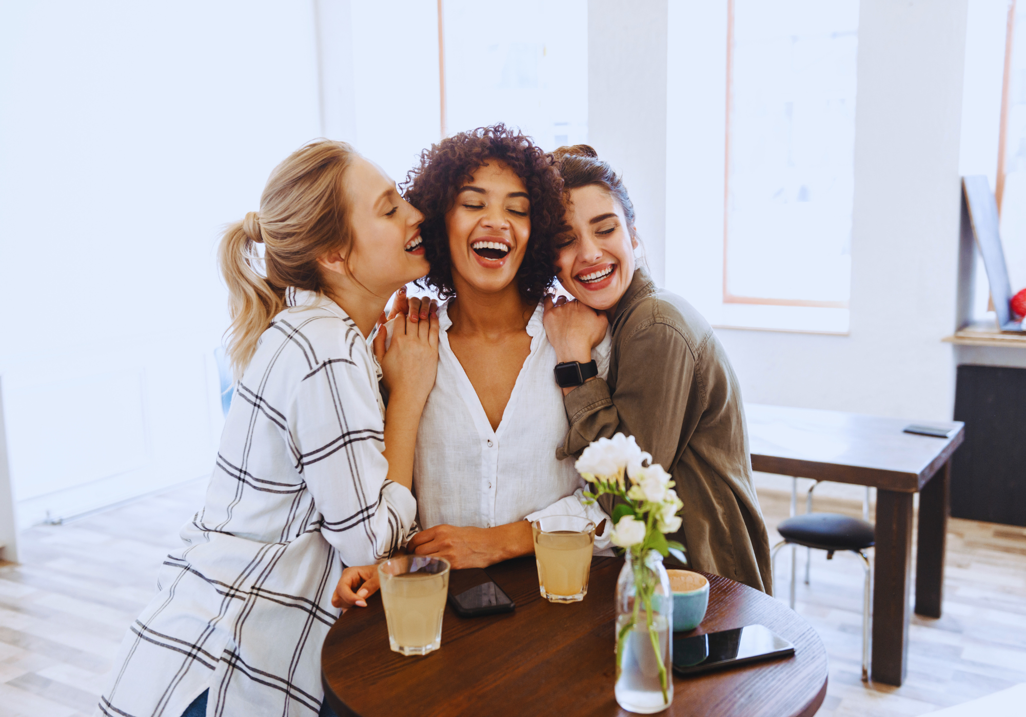 Tres amigas riendo y abrazándose en una cafetería, disfrutando de su compañía.