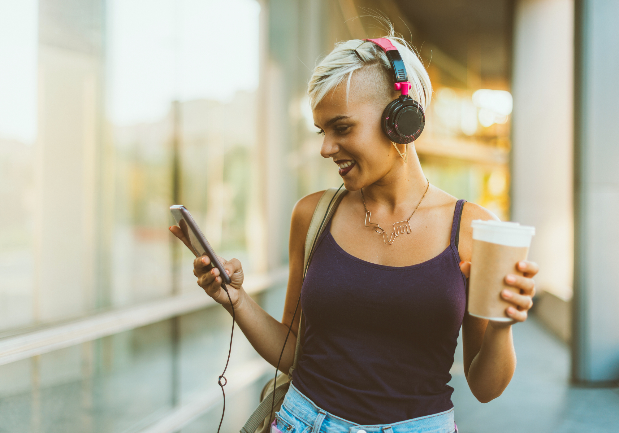 Mujer con audífonos y peinado moderno disfrutando de música mientras sostiene un café y revisa su teléfono.