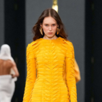 Modelo luce un vestido amarillo con estructura plisada y abertura frontal en el desfile de Balmain en París Fashion Week 2025. La pieza resalta la elegancia arquitectónica característica de la firma.