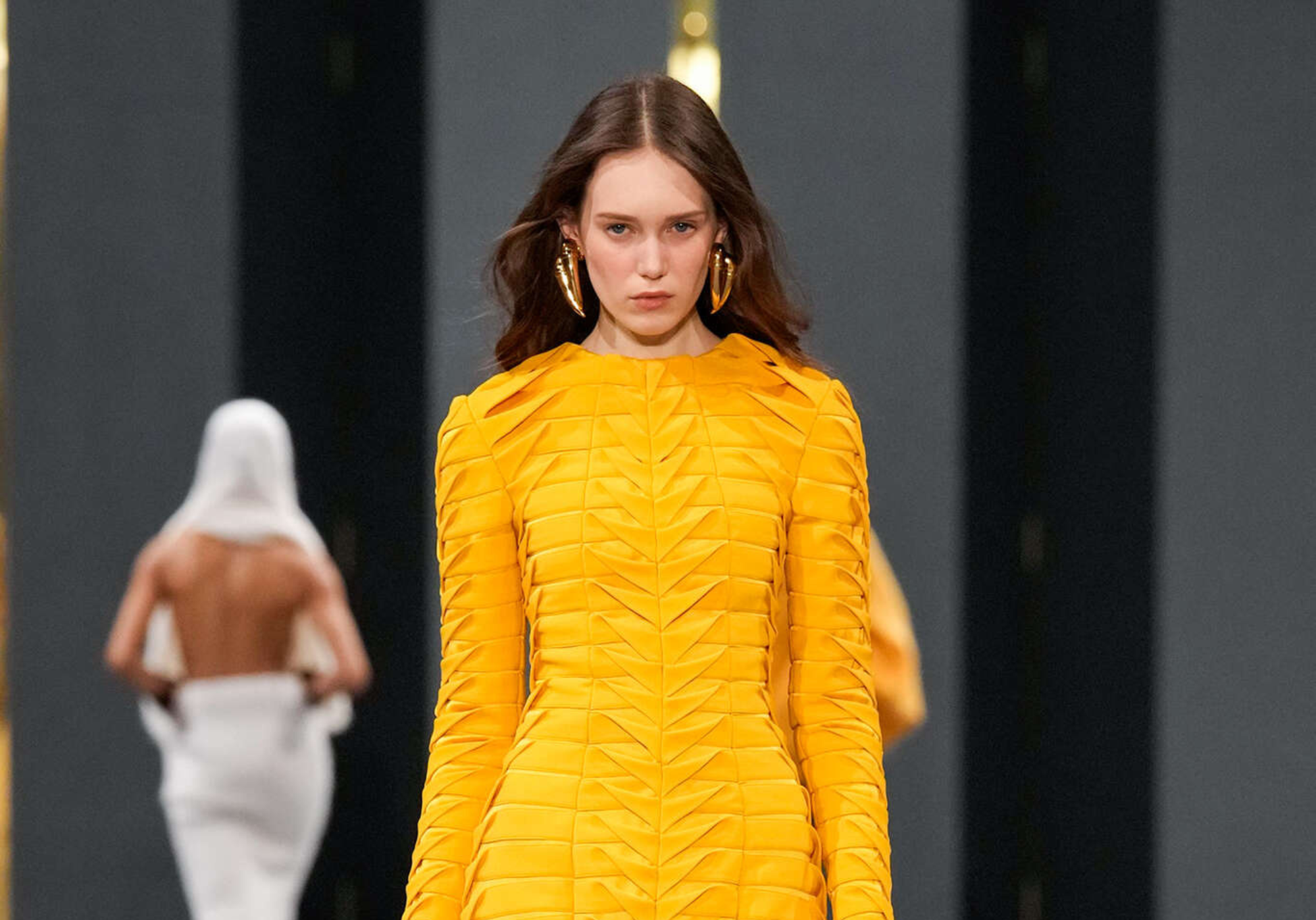 Modelo luce un vestido amarillo con estructura plisada y abertura frontal en el desfile de Balmain en París Fashion Week 2025. La pieza resalta la elegancia arquitectónica característica de la firma.