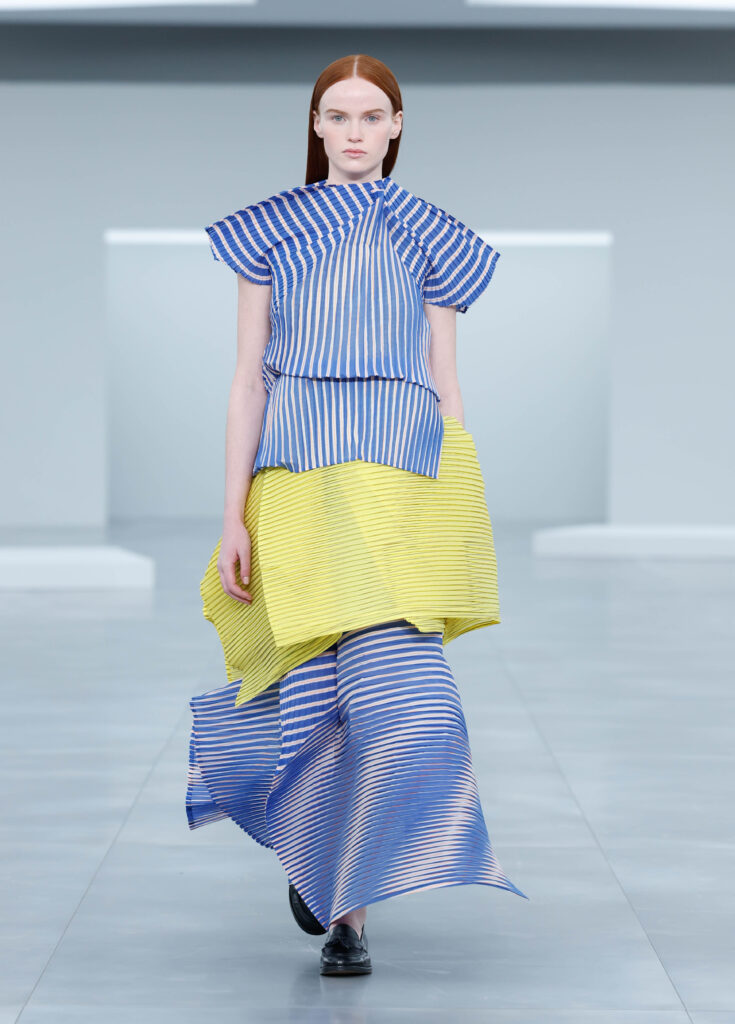  Modelo de Issey Miyake en la Semana de la Moda de París 2025 con un conjunto de líneas estructuradas en azul y amarillo. La silueta se construye a partir de capas asimétricas y plisados geométricos, creando una sensación de movimiento y volumen en cada paso.