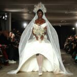 Modelo con un vestido blanco estructurado con velo y corona de flores camina por la pasarela de Weinsanto en la Semana de la Moda de París 2025. El diseño combina teatralidad y opulencia, evocando una estética de realeza moderna.