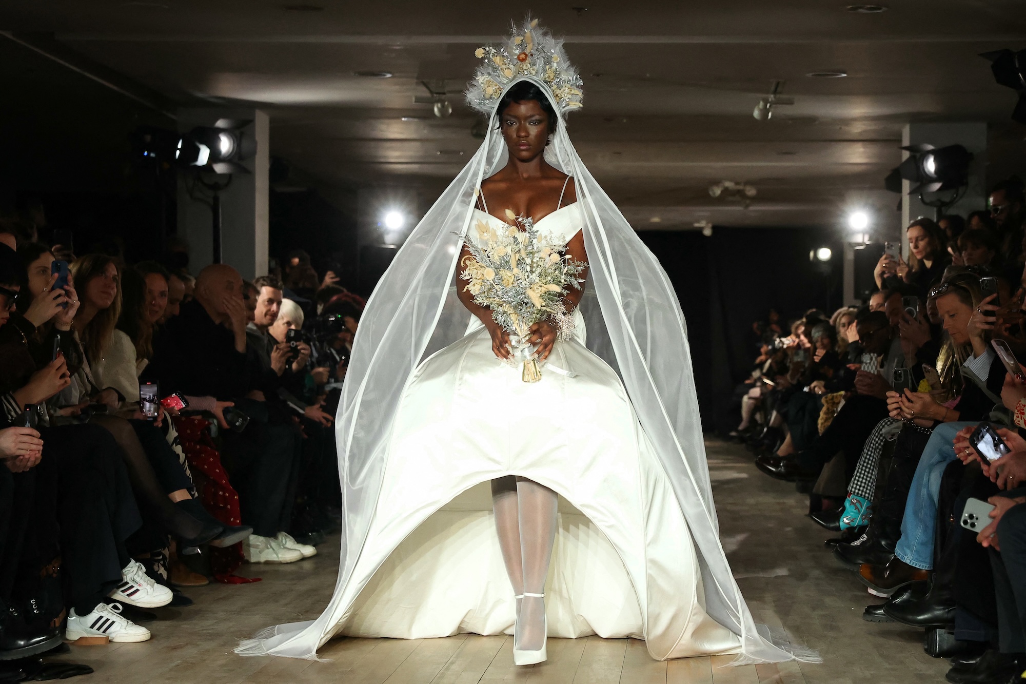 Modelo con un vestido blanco estructurado con velo y corona de flores camina por la pasarela de Weinsanto en la Semana de la Moda de París 2025. El diseño combina teatralidad y opulencia, evocando una estética de realeza moderna.