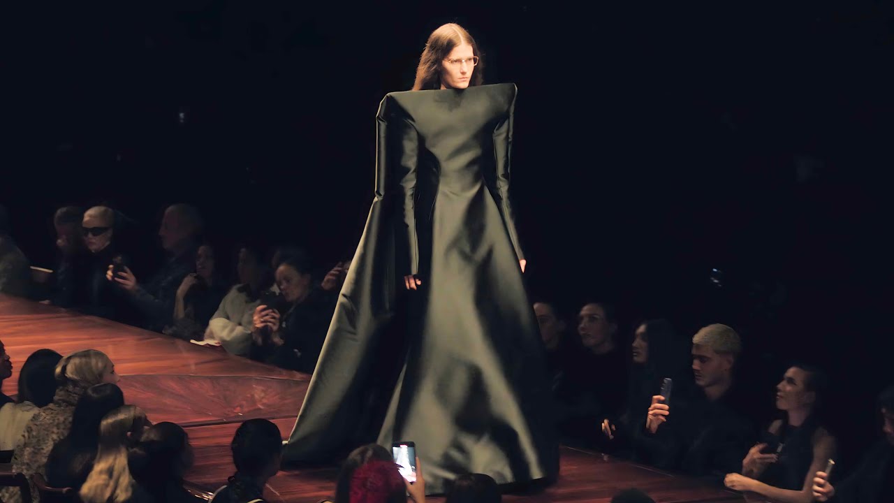 Desfile de Balenciaga en la Semana de la Moda de París 2025, con una modelo luciendo un vestido de líneas vanguardistas y una estética futurista en un ambiente oscuro e impactante.