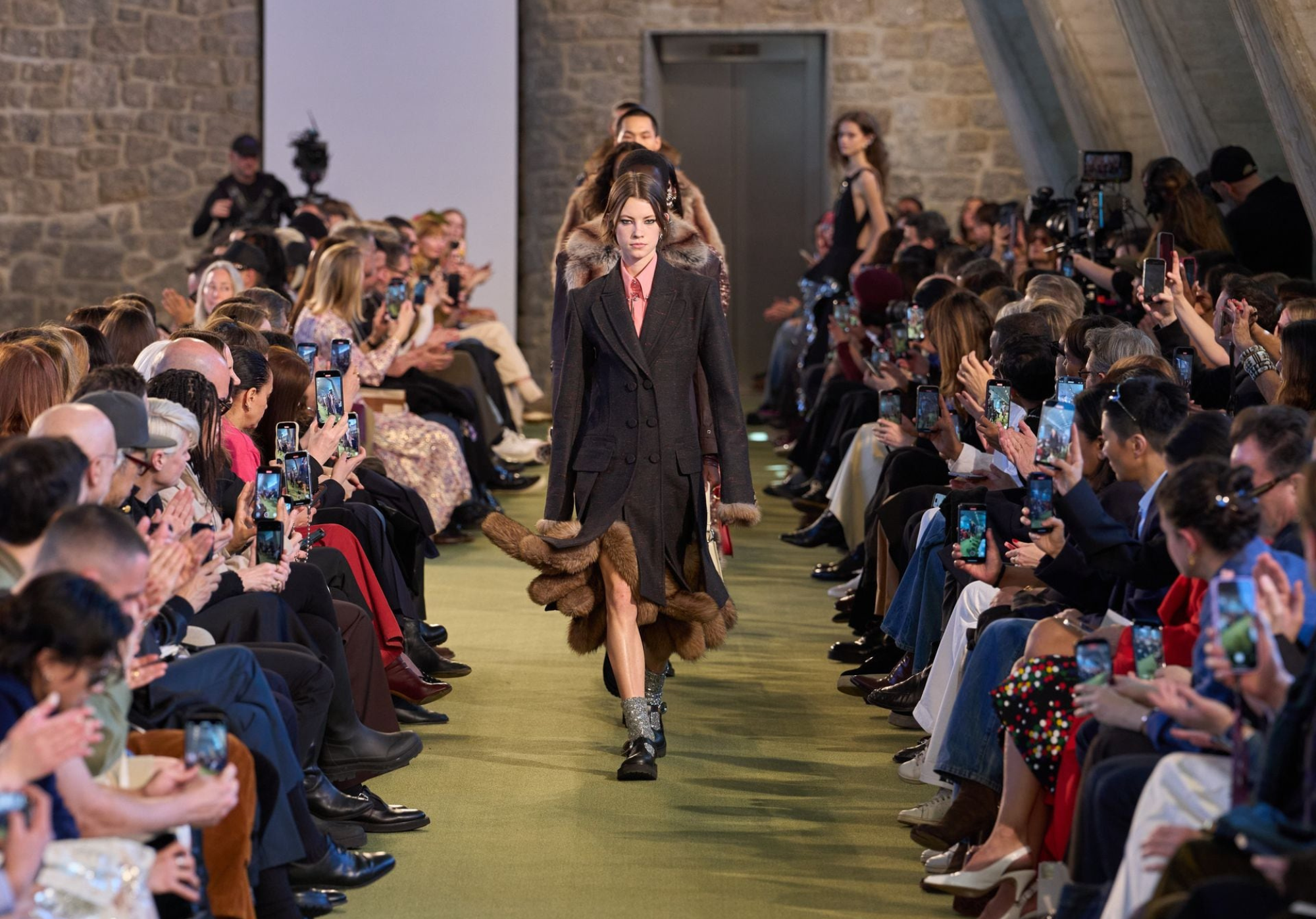 Modelo desfilando en la pasarela de Rabanne en la Semana de la Moda de París, con un abrigo estructurado con detalles de piel, rodeada de un público atento.