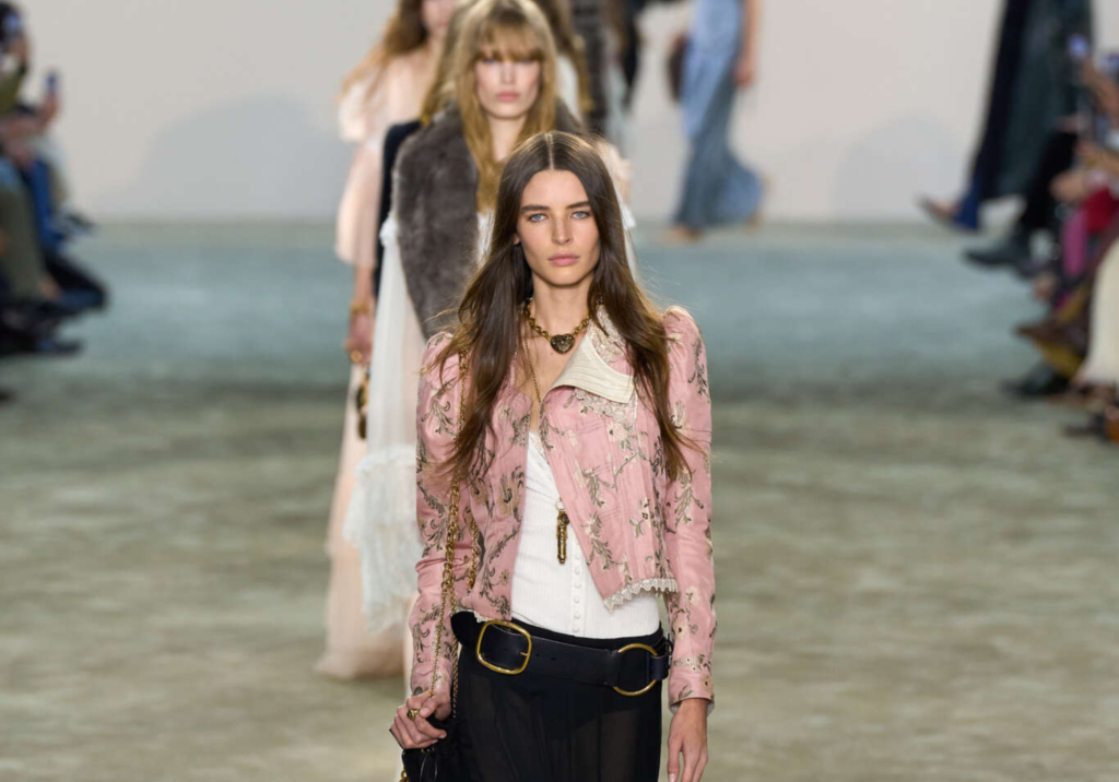 Modelo liderando la pasarela de Chloé con un look bohemio compuesto por una chaqueta rosa bordada, un corsé blanco ajustado y una falda larga negra de gasa semitransparente. Los accesorios dorados y el bolso de cuero negro con cadena refuerzan la esencia vintage y romántica de la colección otoño-invierno 2025.