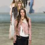 Modelo liderando la pasarela de Chloé con un look bohemio compuesto por una chaqueta rosa bordada, un corsé blanco ajustado y una falda larga negra de gasa semitransparente. Los accesorios dorados y el bolso de cuero negro con cadena refuerzan la esencia vintage y romántica de la colección otoño-invierno 2025.