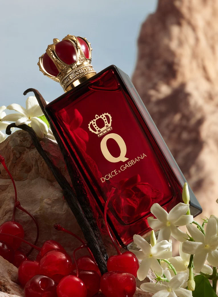 Perfume Q de Dolce & Gabbana en un frasco rojo con corona dorada, rodeado de cerezas y flores blancas.