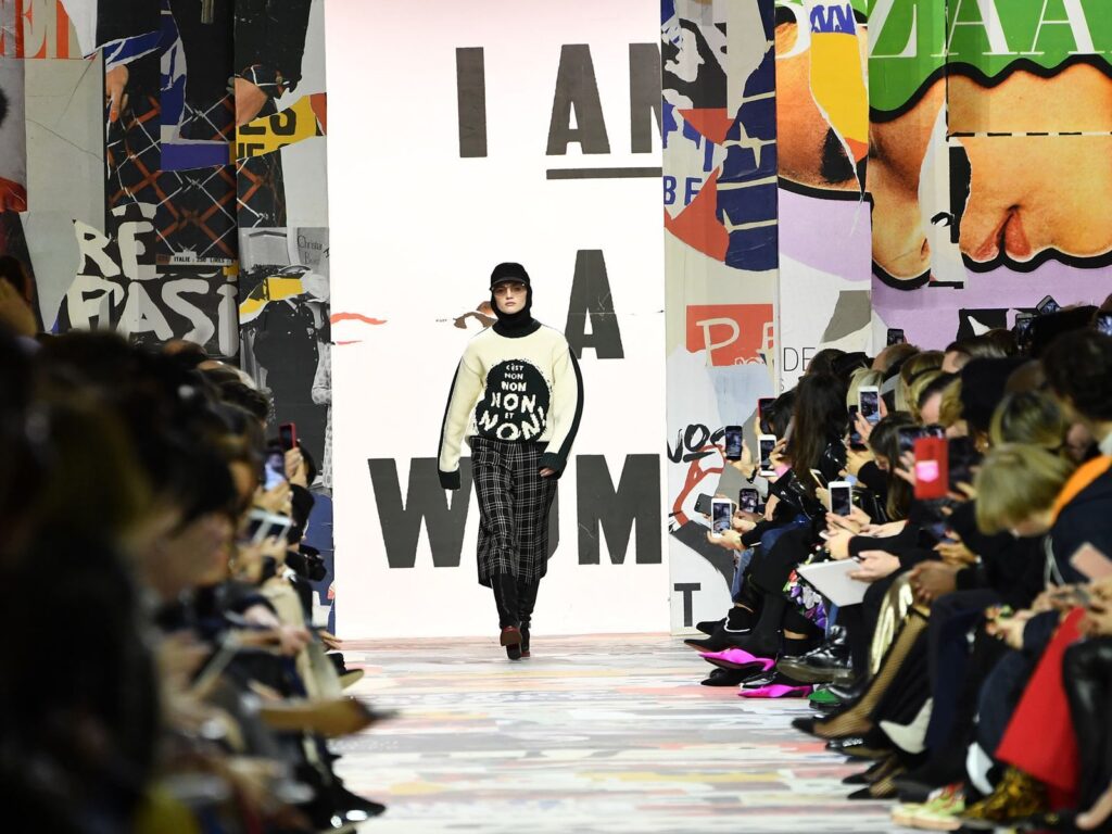 Modelo desfilando en la pasarela de Dior con un suéter de mensaje feminista en un escenario decorado con carteles de protesta y la frase 'I Am a Woman' de fondo. La audiencia captura el momento con sus teléfonos, reflejando cómo la moda y la reivindicación social convergen en la Semana de la Moda de París.