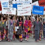 "Desfile de Chanel en la Semana de la Moda de París con modelos marchando en una protesta simbólica por los derechos de las mujeres. Vestidas con trajes coloridos y tweed característico de la firma, sostienen pancartas con mensajes feministas como 'History is Her Story' y 'Women’s Rights Are More Than Alright'. La escena representa la unión entre la moda y la reivindicación social, destacando la pasarela como un espacio de protesta y empoderamiento.