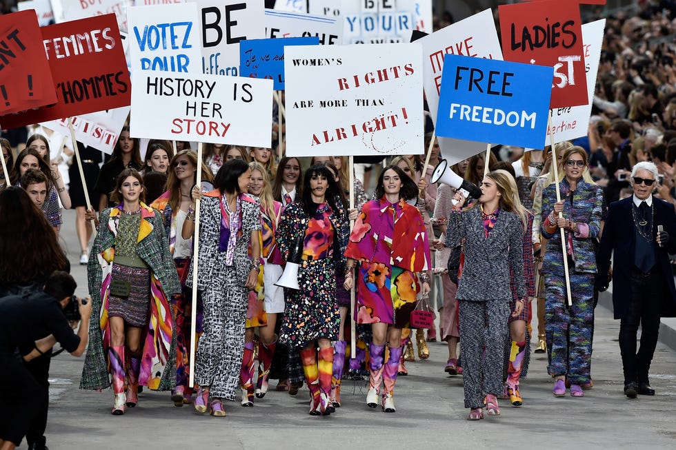 "Desfile de Chanel en la Semana de la Moda de París con modelos marchando en una protesta simbólica por los derechos de las mujeres. Vestidas con trajes coloridos y tweed característico de la firma, sostienen pancartas con mensajes feministas como 'History is Her Story' y 'Women’s Rights Are More Than Alright'. La escena representa la unión entre la moda y la reivindicación social, destacando la pasarela como un espacio de protesta y empoderamiento.