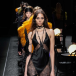 Modelos desfilando en la Semana de la Moda de París 2025 para Nina Ricci. En primer plano, un vestido negro satinado con encaje y transparencias, adornado con detalles de plumas y tiras entrecruzadas en el escote. Detrás, looks con texturas de piel sintética en tonos vibrantes y sedas en colores oscuros, reflejando la estética sensual y dramática de la colección.