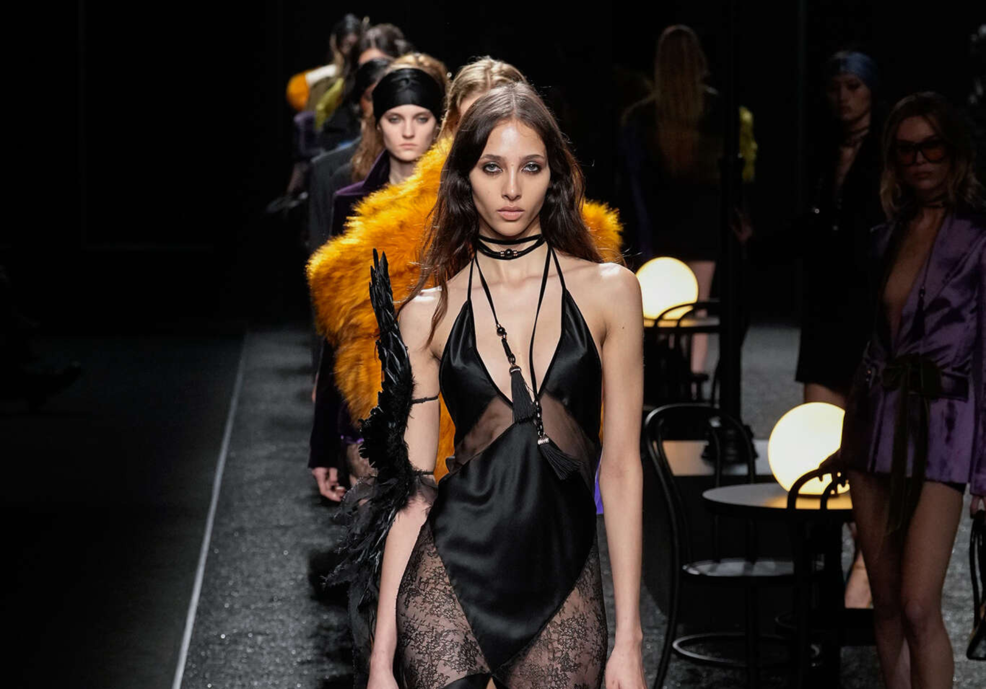 Modelos desfilando en la Semana de la Moda de París 2025 para Nina Ricci. En primer plano, un vestido negro satinado con encaje y transparencias, adornado con detalles de plumas y tiras entrecruzadas en el escote. Detrás, looks con texturas de piel sintética en tonos vibrantes y sedas en colores oscuros, reflejando la estética sensual y dramática de la colección.