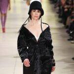 Sarah Paulson desfilando para Miu Miu en París Fashion Week. La actriz luce un abrigo negro satinado con un escote pronunciado y un sombrero tipo cloche, fusionando elegancia clásica con un estilo contemporáneo.