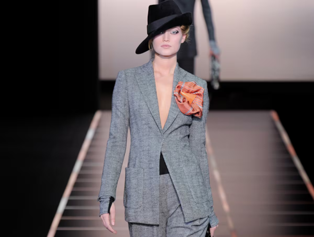 Modelo en la pasarela luciendo un traje sastre gris con solapas marcadas, acompañado de un sombrero negro inclinado y un detalle de tela naranja en el hombro. El look evoca la influencia del icónico Le Smoking de Yves Saint Laurent, representando el empoderamiento femenino y la moda como símbolo de lucha y elegancia.